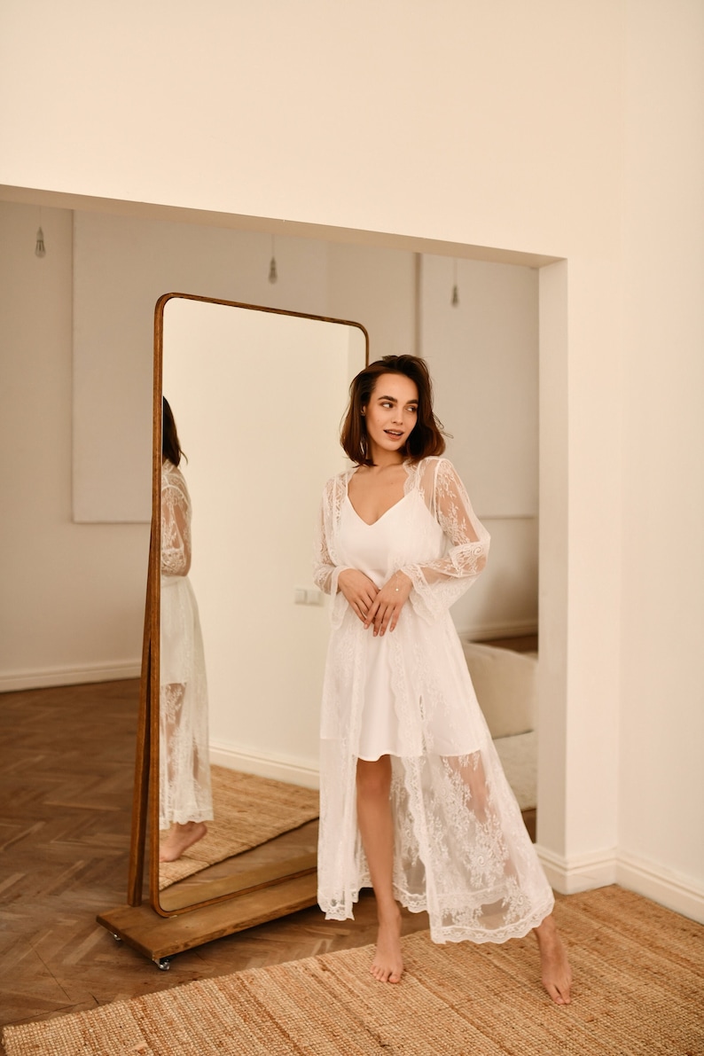 May include: A woman wearing a white lace robe stands in front of a full-length mirror. The robe has long sleeves and a V-neckline. The woman is looking at her reflection in the mirror.