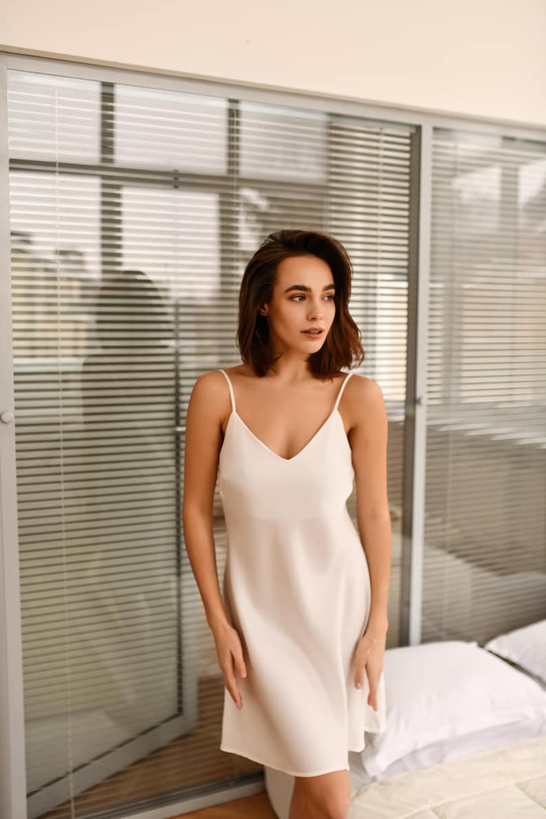 May include: A woman wearing a white slip dress stands in front of a window with blinds. She has short brown hair and is looking to the side.