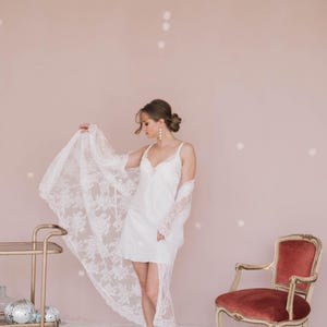 May include: A woman wearing a white lace robe over a white slip dress. She is standing in front of a pink wall with white polka dots. There is a chair with a red velvet seat in the background.