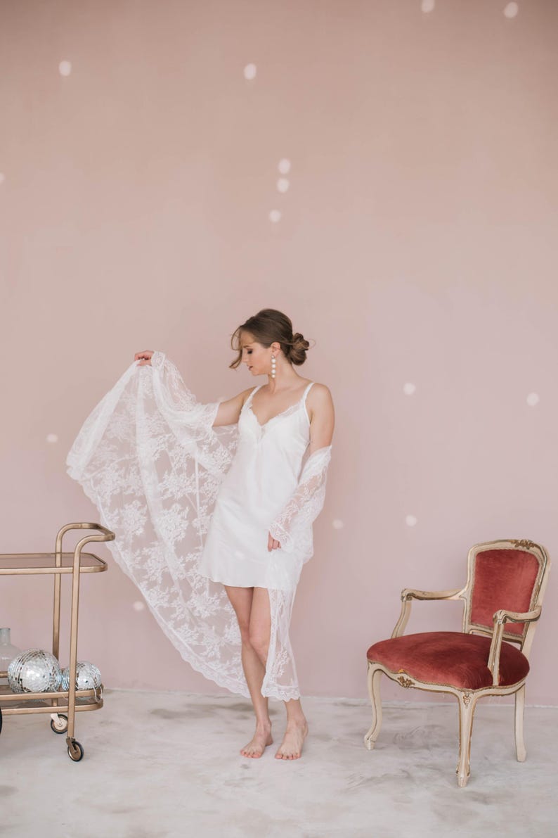 May include: A woman wearing a white lace robe over a white slip dress. She is standing in front of a pink wall with white polka dots. There is a chair with a red velvet seat in the background.