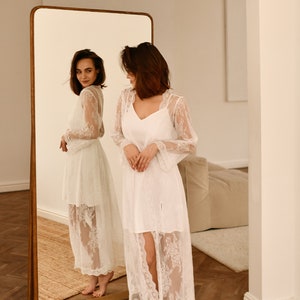 May include: A woman wearing a white lace robe stands in front of a full-length mirror. The robe has long sleeves and a V-neckline. The woman is looking at her reflection in the mirror.