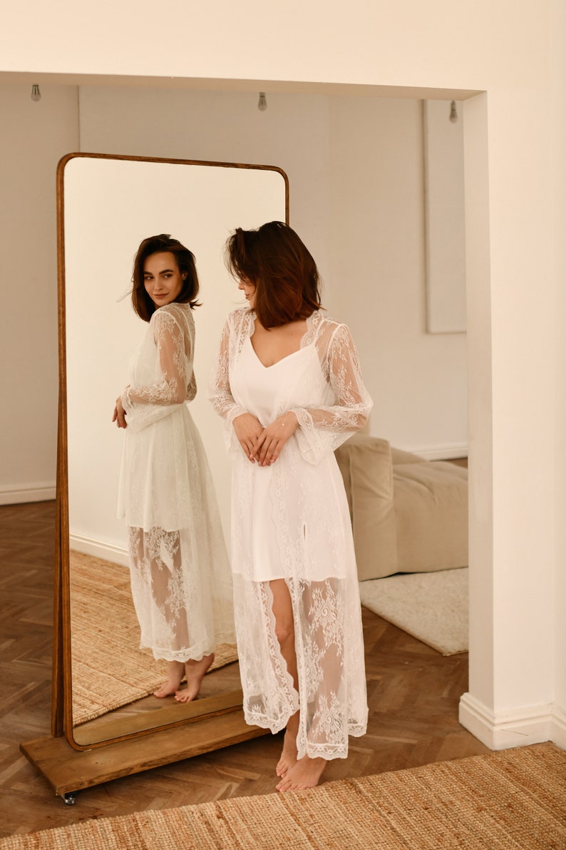 May include: A woman wearing a white lace robe stands in front of a full-length mirror. The robe has long sleeves and a V-neckline. The woman is looking at her reflection in the mirror.