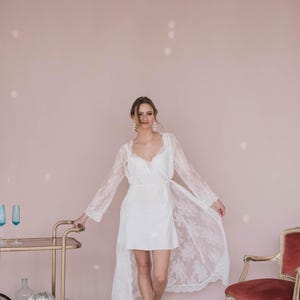 May include: A woman wearing a white lace robe with a white slip dress underneath. She is standing in front of a pink wall with a gold bar cart and a red chair in the background.