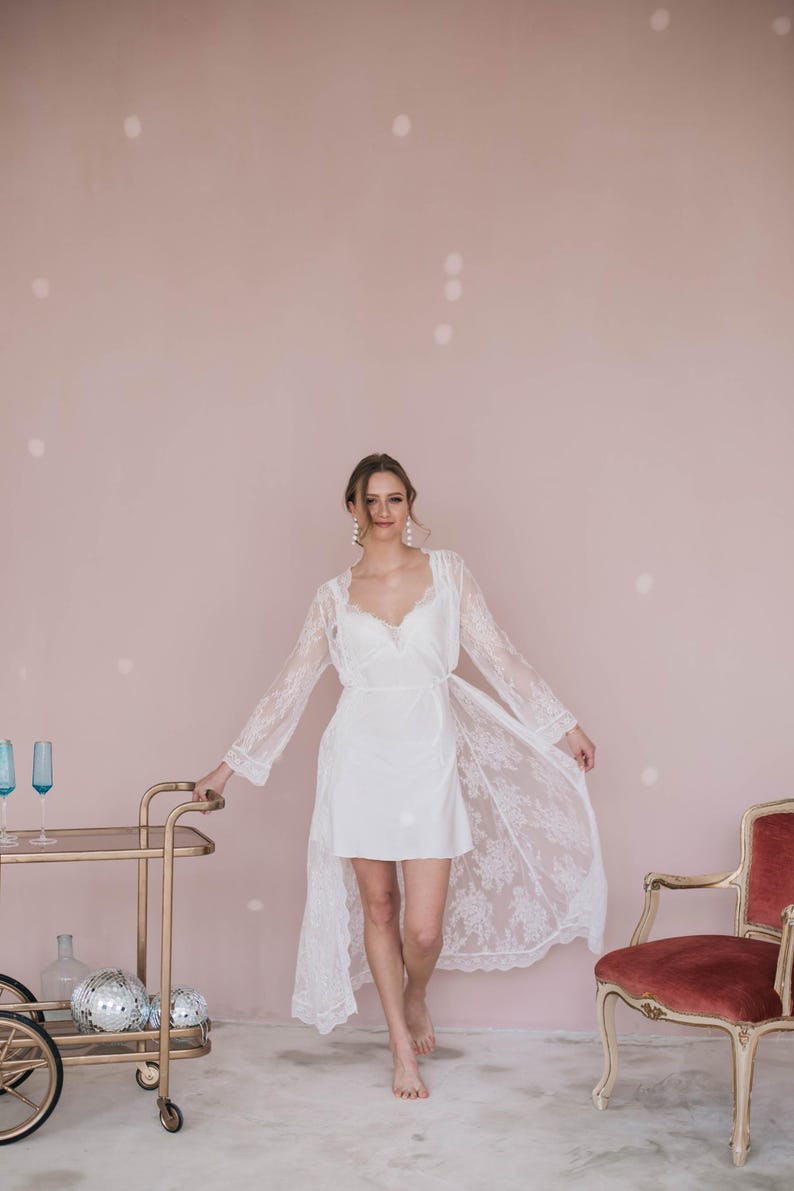 May include: A woman wearing a white lace robe with a white slip dress underneath. She is standing in front of a pink wall with a gold bar cart and a red chair in the background.
