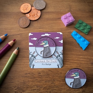 May include: A purple and white enamel pin featuring a cartoon duck with the text "I'm Bread Inside" and "Cormack the Duck Pin Badge".