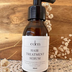 Könnte beinhalten: Eine braune Glasflasche mit einem schwarzen Pumpspender, beschriftet mit "eden HAIR TREATMENT SERUM". Das Etikett besagt auch "HYDRATING OILS FOR A PRE-WASH TREATMENT. SUITABLE FOR DRY & BLEACHED HAIR" und "100ml". Die Flasche steht auf einer Holzoberfläche.