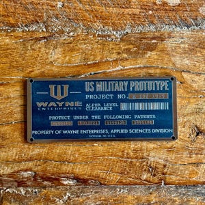 May include: A rectangular, dark blue metal plaque with the text "US MILITARY PROTOTYPE" and "WAYNE ENTERPRISES". The plaque includes a project number, patent numbers, and the text "PROPERTY OF WAYNE ENTERPRISES, APPLIED SCIENCES DIVISION".