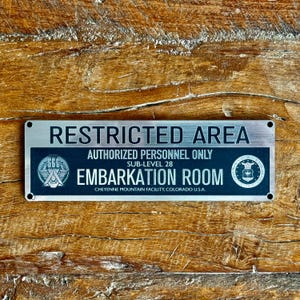 Op de afbeelding: Een rechthoekig metalen bord met de woorden "RESTRICTED AREA" in hoofdletters. Daaronder staat "AUTHORIZED PERSONNEL ONLY" en "EMBARKATION ROOM". Het bord bevat ook de tekst "CHEYENNE MOUNTAIN FACILITY, COLORADO U.S.A."