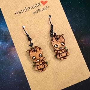 May include: Wooden dangle earrings featuring a cartoon character design. The earrings are brown with black accents and green details. The earrings are displayed on a brown card with the text "Handmade with love".