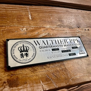 May include: A silver metal plaque with black text, including "WALTHER PPK" and "QUARTERMASTER BRANCH ISSUED." The plaque features the text "ROYAL SECRET INTELLIGENCE SERVICE" and "AGENT ASSIGNED 007." The plaque has a black border and is rectangular in shape.