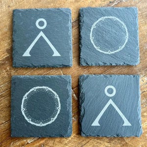 May include: Four square slate coasters with white geometric designs. Two coasters feature a triangle with a circle above, and the other two have a circular design with intricate patterns. The coasters are dark gray.