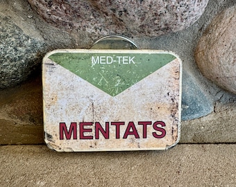May include: Rectangular, distressed metal sign with the text "MED-TEK" at the top and "MENTATS" in large, maroon letters below. The sign has a white and green chevron design and a rusty, aged appearance.