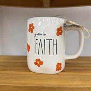 May include: A white ceramic mug with a rounded handle and a tag attached. The mug features orange flower illustrations and the black text "grow in FAITH" printed on the front. The design is simple with a smooth finish and a sturdy, curved handle.