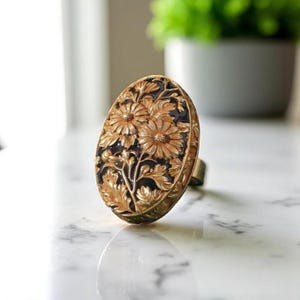 Può includere: Anello ovale color oro con un design floreale. L'anello presenta un motivo in rilievo di fiori e foglie su uno sfondo scuro. L'anello è appoggiato su una superficie di marmo.