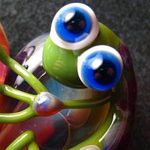 May include: A glass figurine of a green frog with large blue eyes and white pupils. The frog is sitting on a multi-coloured base.