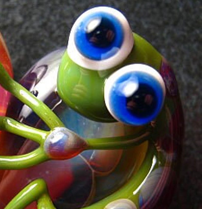 May include: A glass figurine of a green frog with large blue eyes and white pupils. The frog is sitting on a multi-coloured base.