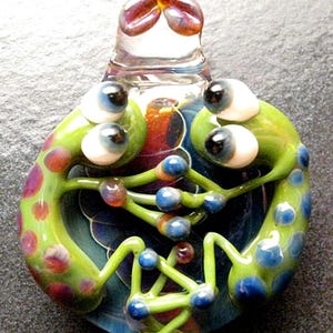 May include: A glass pendant featuring two green frogs with blue and red spots, intertwined with their legs and facing each other. The frogs have large white eyes with black pupils and are set against a swirling background of blue, red, and green.