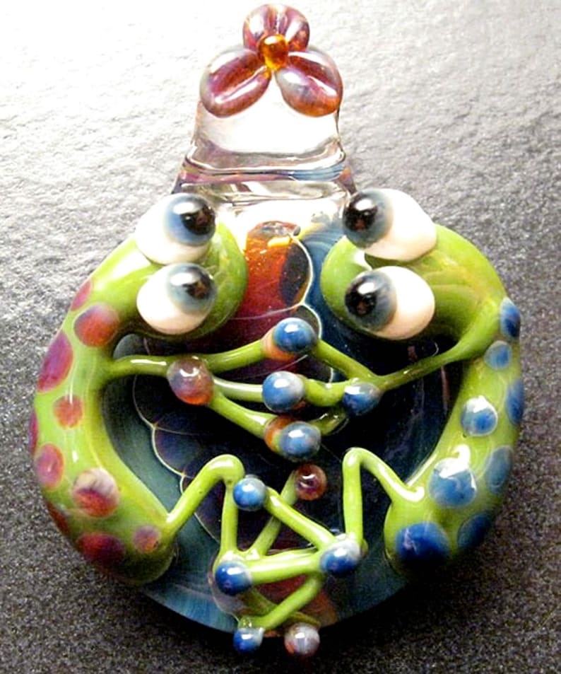 May include: A glass pendant featuring two green frogs with blue and red spots, intertwined with their legs and facing each other. The frogs have large white eyes with black pupils and are set against a swirling background of blue, red, and green.