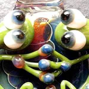 May include: A glass sculpture of two green frogs with large, colorful eyes. The frogs are intertwined and have a swirling, multi-colored background.