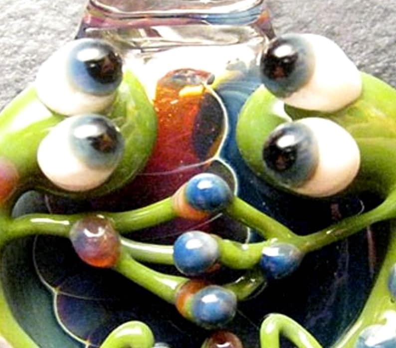 May include: A glass sculpture of two green frogs with large, colorful eyes. The frogs are intertwined and have a swirling, multi-colored background.