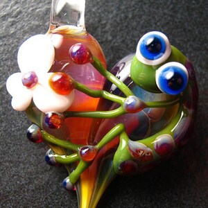May include: A glass pendant with a heart-shaped design featuring a whimsical green creature with large blue eyes, surrounded by white flowers and green stems. The pendant has a mix of red, orange, yellow, and purple glass accents.