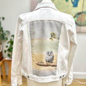 May include: White denim jacket with a patch on the back featuring a gray owl perched on a branch with a green leafy background.