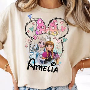 Könnte beinhalten: Beiges T-Shirt mit einem Minnie-Mouse-inspirierten Design mit den Figuren aus dem Film Frozen. Das Design umfasst eine rosa Schleife, Sterne und den Namen "Amelia" in schwarzer Schrift. Das Shirt ist aus weichem Material.
