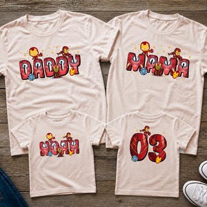 May include: Four light pink t-shirts with matching designs. Two shirts say "DADDY" and "MAMA" in red and black block letters with Iron Man graphics. The other two shirts say "NOAH" and "03" with similar graphics.