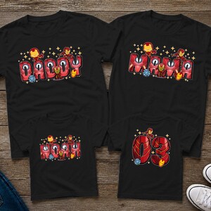 May include: Four black t-shirts with Iron Man-themed designs. Two shirts say "Daddy" and "Mama" in red bubble letters with Iron Man graphics. The other two shirts have "03" and "Mom" in similar styles. Family matching apparel.
