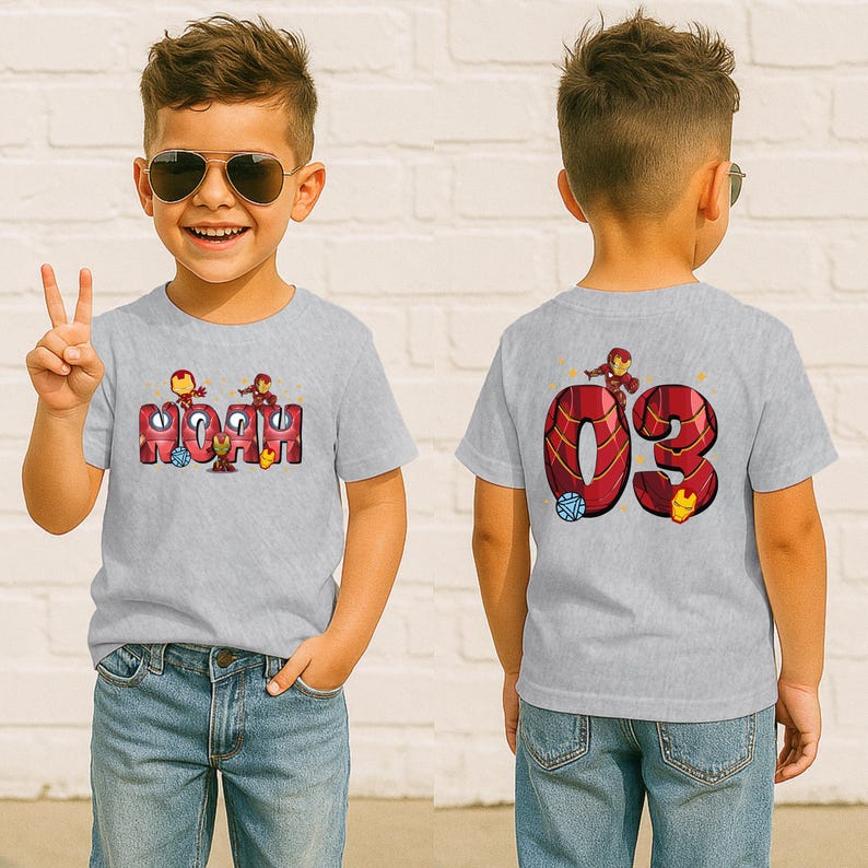 May include: A light gray t-shirt featuring the name "Noah" in a red and gold Iron Man design on the front. The back of the shirt displays the number "03" in a similar design, with Iron Man graphics. The child is wearing blue jeans.