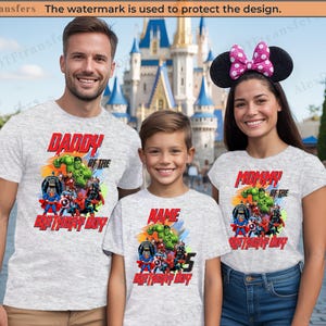 以下が含まれることがあります： スーパーヒーローのグラフィックがプリントされた、お揃いのグレーのTシャツを着た3人。シャツには「Daddy of the Birthday Boy」、「Mommy of the Birthday Boy」、そして「Name Birthday Boy」と書かれています。背景にはお城が写っています。