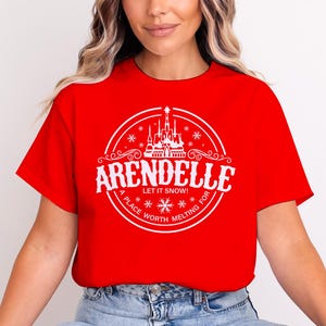 Op de afbeelding: Rood T-shirt met een wit cirkelvormig ontwerp. Het ontwerp bevat het woord "Arendelle", de zin "Let it snow!", een kasteelillustratie en de tekst "A place worth melting for".