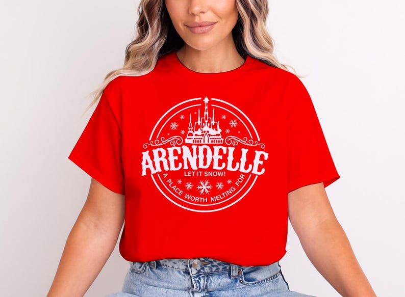Op de afbeelding: Rood T-shirt met een wit cirkelvormig ontwerp. Het ontwerp bevat het woord "Arendelle", de zin "Let it snow!", een kasteelillustratie en de tekst "A place worth melting for".