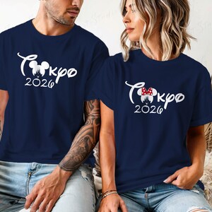 Puede incluir: Camisetas azul marino con texto gr&aacute;fico blanco "Tokyo 2026". Una camiseta presenta un lazo de Minnie Mouse. Las camisetas son de manga corta y las llevan dos personas.