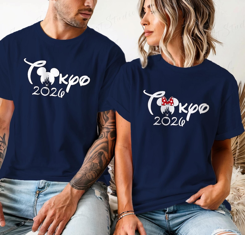 Puede incluir: Camisetas azul marino con texto gr&aacute;fico blanco "Tokyo 2026". Una camiseta presenta un lazo de Minnie Mouse. Las camisetas son de manga corta y las llevan dos personas.