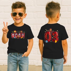 May include: Black t-shirt featuring the name "NOAH" in red and gold letters with Iron Man graphics. The back of the shirt displays the number "03" with Iron Man and other graphics. The boy is wearing blue jeans.
