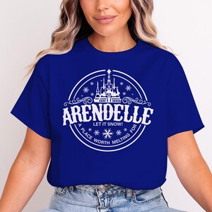 Op de afbeelding: Koningsblauw T-shirt met een wit cirkelvormig ontwerp. Het ontwerp bevat het woord "ARENDELLE", de zin "LET IT SNOW!" en "A PLACE WORTH MELTING FOR". Een kasteel en sneeuwvlokken zijn ook inbegrepen.