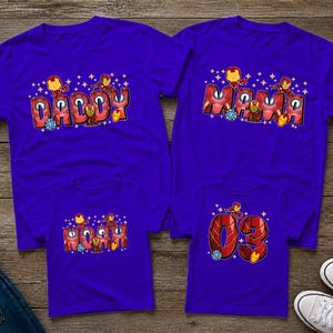 May include: Four purple t-shirts with Iron Man-themed designs. Two shirts say "Daddy" and "Mama" in red bubble letters, with Iron Man figures. The other two shirts have "Mama" and the number "03" in a similar style.