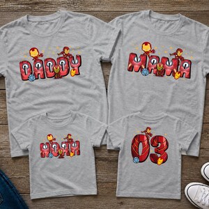 May include: Four gray t-shirts with matching designs. The shirts feature the words "Daddy," "Mama," "Noah," and "03" in red and orange lettering, with cartoon character graphics. The shirts are displayed on a wooden surface.