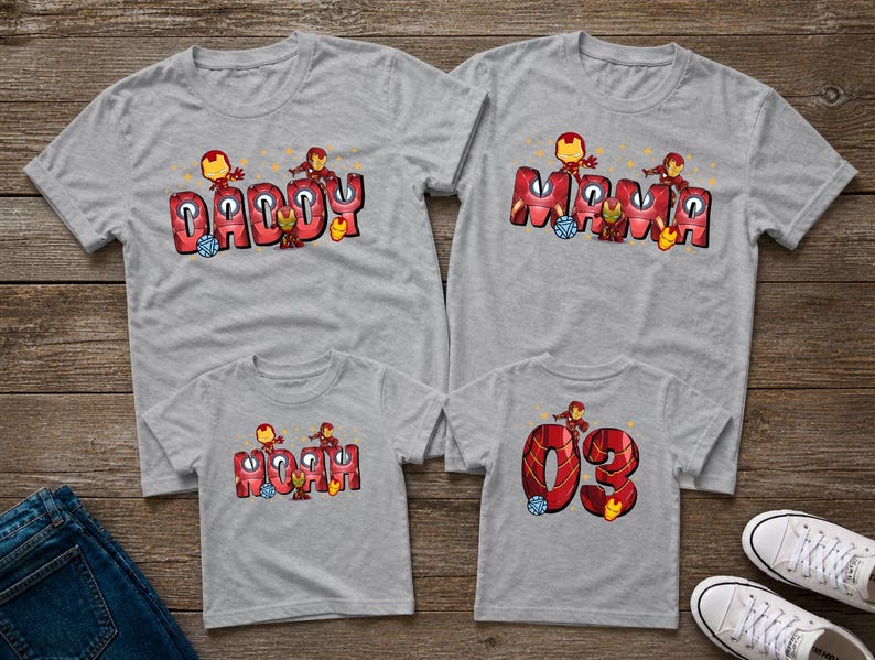 May include: Four gray t-shirts with matching designs. The shirts feature the words "Daddy," "Mama," "Noah," and "03" in red and orange lettering, with cartoon character graphics. The shirts are displayed on a wooden surface.