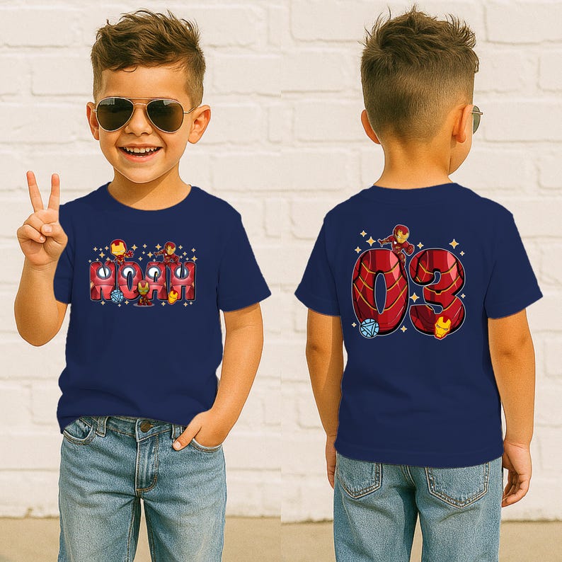 May include: Navy blue t-shirt featuring the name "Noah" in a red and gold Iron Man design on the front. The back of the shirt displays the number "03" with Iron Man graphics. The shirt is worn by a child.
