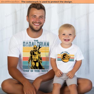 K&ouml;nnte beinhalten: Passende wei&szlig;e T-Shirts mit Retro-Design. Ein Shirt sagt "The Dadalorian" mit einem Mandalorianer-Grafik, das andere sagt "The Child" mit einer Grogu-Grafik. Beide Shirts haben einen gestreiften Hintergrund.