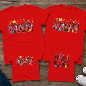 May include: Four red t-shirts with superhero-themed designs. Two shirts say "Daddy" and "Momma" in a stylized font, and the other two shirts have the number "03". The shirts are displayed on a wooden surface.