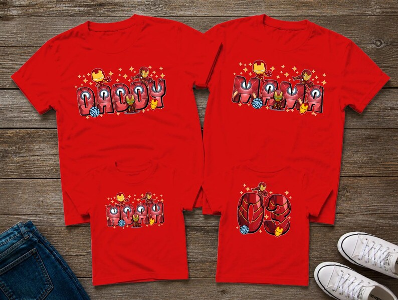May include: Four red t-shirts with superhero-themed designs. Two shirts say "Daddy" and "Momma" in a stylized font, and the other two shirts have the number "03". The shirts are displayed on a wooden surface.