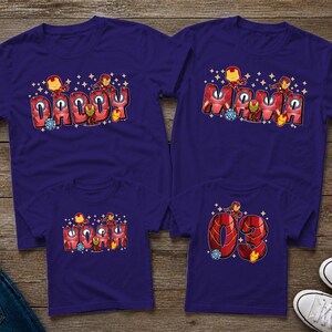 May include: Four matching navy blue t-shirts with Iron Man-themed designs. Two adult shirts say "Daddy" and "Mama" in red bubble letters. The other two shirts say "03" and "Mini" in red bubble letters. All shirts have Iron Man graphics.
