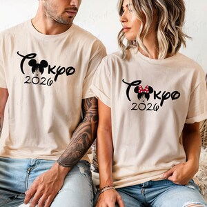 Puede incluir: Dos camisetas beige con texto y gr&aacute;ficos negros. Las camisetas presentan la palabra "Tokyo" en cursiva, una silueta de Mickey Mouse, la palabra "2026" y un gr&aacute;fico de castillo. Una camiseta tiene un lazo rojo.