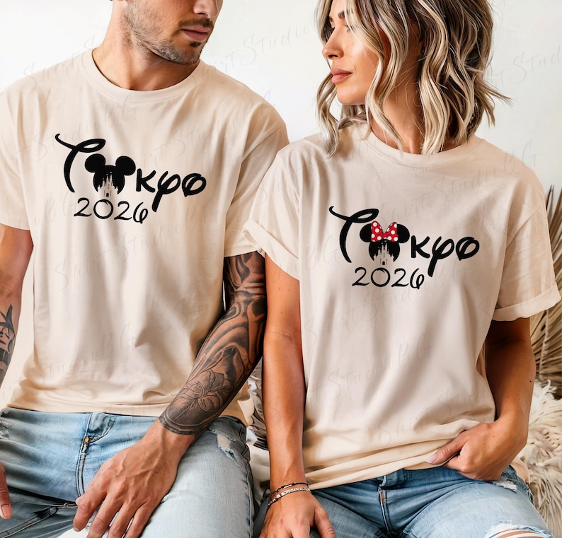 Puede incluir: Dos camisetas beige con texto y gr&aacute;ficos negros. Las camisetas presentan la palabra "Tokyo" en cursiva, una silueta de Mickey Mouse, la palabra "2026" y un gr&aacute;fico de castillo. Una camiseta tiene un lazo rojo.
