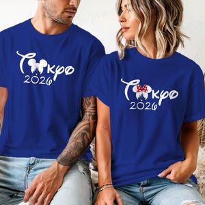 Puede incluir: Dos camisetas azul real con texto blanco "Tokyo 2026" y un dise&ntilde;o inspirado en Mickey Mouse. Una camiseta tiene un lazo de Minnie Mouse. Las camisetas son de manga corta.