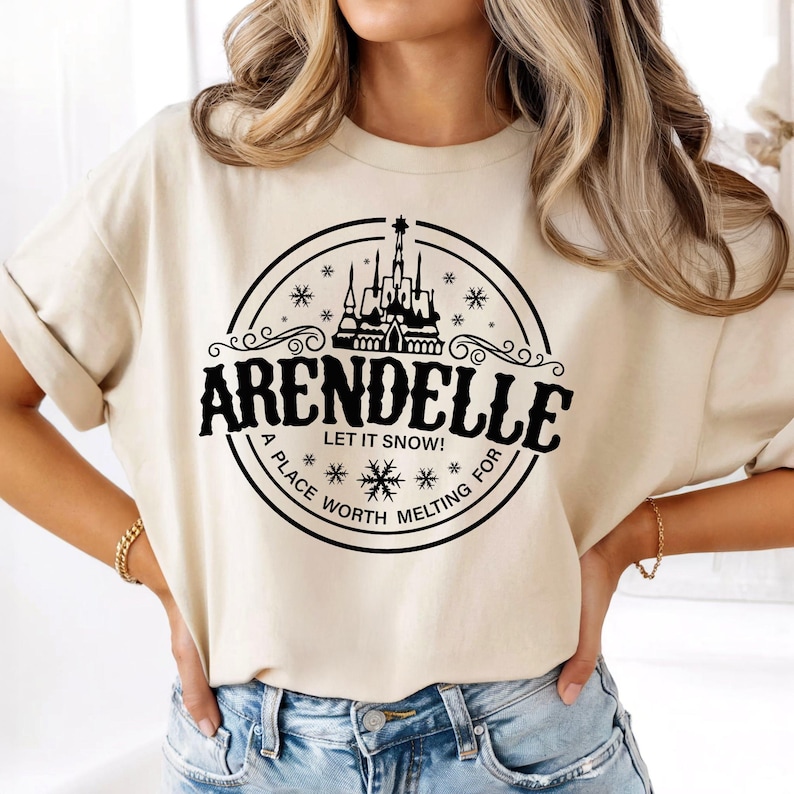 Op de afbeelding: Beige t-shirt met een zwart cirkelvormig ontwerp met een kasteelsilhouet, sneeuwvlokken en het woord "Arendelle". De tekst "LET IT SNOW!" en "A PLACE WORTH MELTING FOR" zijn ook inbegrepen.