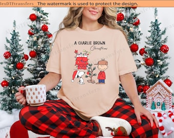 May include: A tan t-shirt with the text "A Charlie Brown Christmas" and a graphic of Charlie Brown, Snoopy, and a Christmas tree. The person is wearing red and black plaid pajama pants and holding a mug with a gingerbread man design.